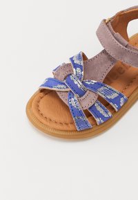 Lilla semskede sandaler med en blå, blomsterprydd rem og et knyttet design. Brun teksturert innersåle og en fleksibel gummisåle.