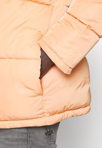 Peach-colored puffer jacket with quilted sections, featuring a side pocket and a cinched hem. Fabric appears smooth and water-resistant.