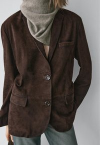 Brązowa zamszowa marynarka z dwoma przednimi kieszeniami, jedną kieszenią na piersi i dwoma guzikami. Nosi się ją na beżowym szaliku i łączy z dżinsami.
