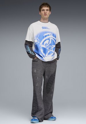 Junger Mann trägt ein weiß-blaues Langarmshirt mit "Fast & Furious"-Grafik, graue strukturierte Hosen mit Reifenprofilmustern und blaue Turnschuhe.