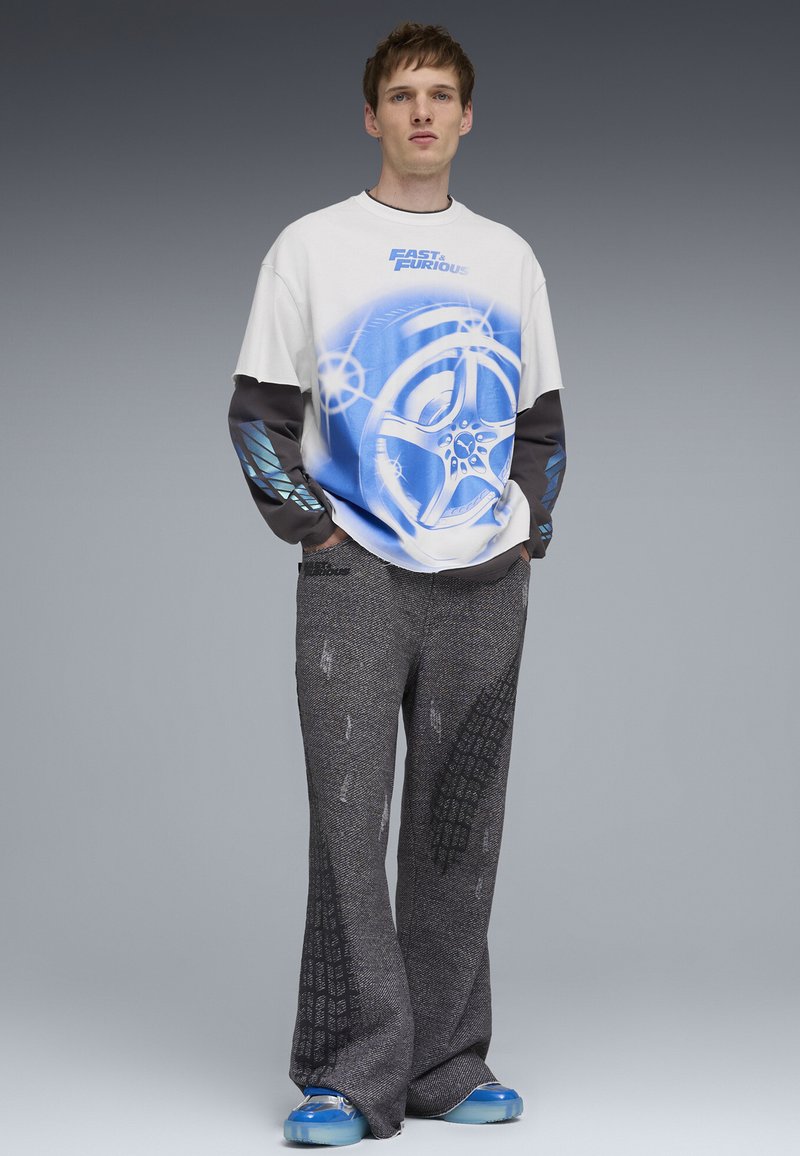 Junger Mann trägt ein weiß-blaues Langarmshirt mit "Fast & Furious"-Grafik, graue strukturierte Hosen mit Reifenprofilmustern und blaue Turnschuhe.