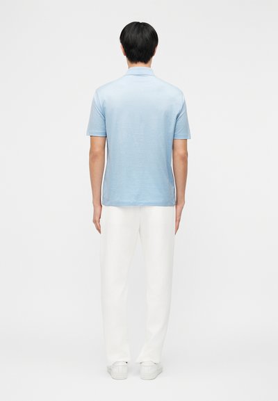 Άνδρας με την πλάτη προς τα εμάς φοράει ανοιχτό μπλε κοντομάνικο πουκάμισο, λευκό παντελόνι και λευκά αθλητικά παπούτσια μπροστά σε απλό ανοιχτό φόντο.