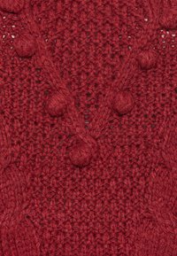 Maglione rosso a maglia con un motivo testurizzato, con accenti di pompom in rilievo e scollatura a V. Il tessuto appare spesso e caldo.