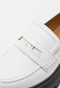 Mocassin en cuir blanc avec un design cousu, mettant en valeur un accent métal doré et une semelle noire texturée. Finition lisse avec des détails subtils.