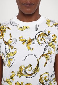 T-shirt bianco in cotone con motivo barocco giallo e nero, scollo tondo e maniche corte. Presenta il logo del marchio integrato nel design.