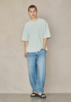 Junger Mann steht vor einer neutralen Wand, trägt ein helles, übergroßes T-Shirt, weite blaue Jeans und schwarze Sandalen.