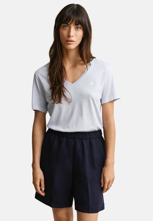 Frau mit langen braunen Haaren, die ein hellgraues T-Shirt mit V-Ausschnitt und dunkelblaue, hoch taillierte Shorts trägt, stehend vor einem weißen Hintergrund.
