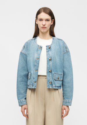 Kobieta z prostymi brązowymi włosami, ubrana w jasnoniebieską zapinaną na guziki kurtkę dżinsową, biały top i beżowe, szerokie spodnie z wysokim stanem.