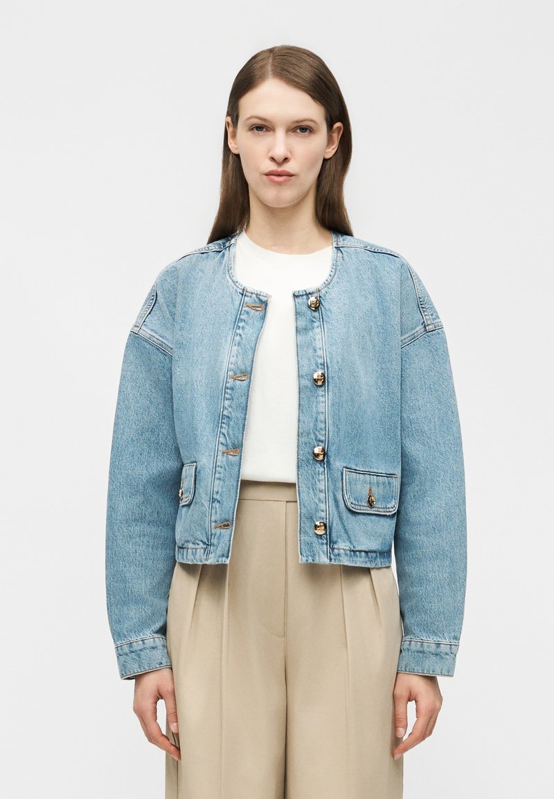 Γυναίκα με ίσια καστανά μαλλιά, που φοράει ένα ανοιχτό μπλε τζιν μπουφάν με κουμπιά, λευκό τοπ και μπεζ παντελόνι με φαρδιά μπατζάκια και ψηλή μέση.