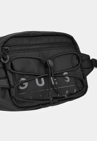 Sac de taille en nylon noir avec une surface texturée, compartiment zippé, fermeture à cordon et détail de logo embossé.