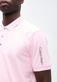 Άνδρας που φοράει ανοιχτό ροζ κοντομάνικο πόλο πουκάμισο με τρία κουμπιά και το λογότυπο "Calvin Klein" τυπωμένο κάθετα στο μανίκι.