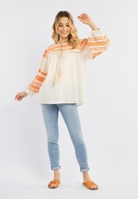 Hvid let bluse med orange geometrisk broderi, lange puffærmer og en snoretræk-udskæring, parret med lyseblå jeans og orange sandaler.