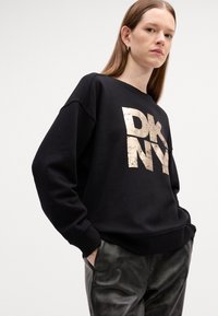 Svart sweatshirt med bred rund hals og nedfalne skuldre. Har metallisk gull "DKNY" logo. Kombinert med blanke svarte bukser.