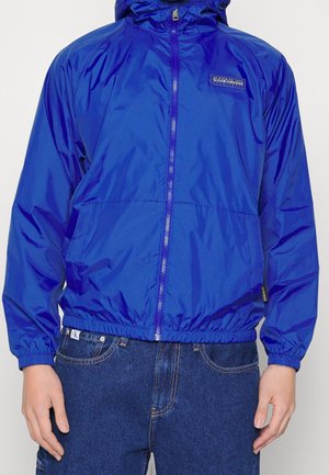 Niebieska kurtka z kapturem na zamek, noszona na ciemnoniebieskich dżinsach z widocznymi metkami marki na piersiach kurtki i w pasie dżinsów.