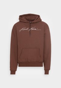 Brun hoodie med dragsko i huvan, känguruficka och vit logotext på framsidan. Tillverkad av mjukt tyg med ribbade ärmslut och nederkant.