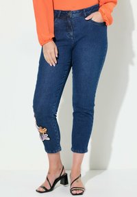 Sininen farkkuhousut, joissa on mukautuva muotoilu, vasemmassa nilkassa koristeellinen kukkakuvio ja keskipitkä vyötärö.