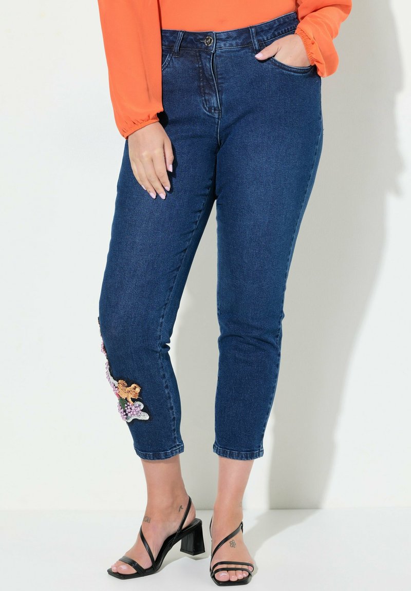 Sininen farkkuhousut, joissa on mukautuva muotoilu, vasemmassa nilkassa koristeellinen kukkakuvio ja keskipitkä vyötärö.