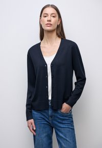 Granatowy sweter zapinany na guziki, wykonany z miękkiego materiału, z dekoltem w kształcie litery V, długimi rękawami i ściągaczami przy mankietach. Nosi się go na białej bluzce i niebieskich dżinsach.