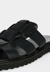 Sandale slide en cuir noir avec trois larges brides et une semelle épaisse texturée, montrant un gros plan des détails avant et latéraux.