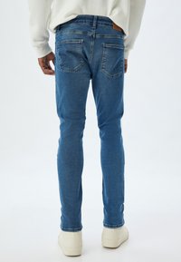 Jeans de mezclilla azul de corte slim con un lavado desvanecido, que cuentan con dos bolsillos traseros y un parche de cuero marrón en la cintura. Diseño clásico con detalles mínimos.