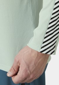 Lichtgroen textuur langsliepend shirt met een zwart-wit gestreepte mouwaccent. Close-up van een hand die het shirt bij de zoom vasthoudt.