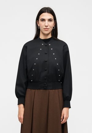 Kobieta z długimi ciemnymi włosami nosi czarną zapinaną kurtkę z oczkami oraz brązową plisowaną spódnicę na jednolitym tle.