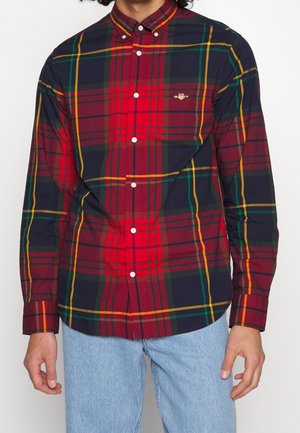 Mężczyzna ubrany w czerwoną, granatową i zieloną koszulę w kratę z długim rękawem i białymi guzikami, do tego jasnoniebieskie dżinsy, stoi na białym tle.