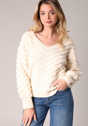 Pull crème épais avec un col en V, présentant un motif en zigzag texturé et des poignets côtelés, associé à un jean en denim bleu clair.