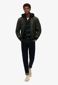 Groene bomberjack over een geruit shirt, donkere jeans en beige sneakers. Heeft ribgebreide boorden, zipzakken en een capuchonlaag eronder.