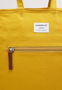 Borsa in tela gialla con una tasca anteriore con zip, un tira in pelle marrone e un'etichetta bianca che mostra il nome e i dettagli del marchio nella parte superiore.