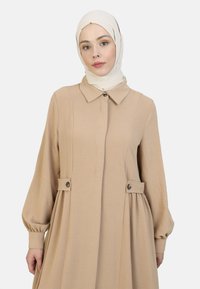 Beige langærmet kjole med krave, knapdetaljer ved taljen og en afslappet, let silhuet. Tekstureret stofoverflade.