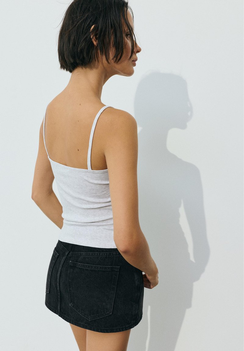 Femme aux cheveux courts et foncés, portant un débardeur blanc et une jupe noire, debout de profil contre un mur clair et uni.