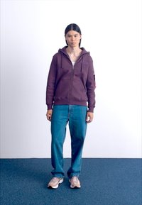 Felpa viola con zip e coulisse, indossata sopra una canotta bianca. Abbinata a jeans larghi blu e sneaker beige. Design semplice e casual.