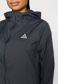 Ελαφρύ, ναυτικό μπουφάν αδιάβροχο με φερμουάρ μπροστά, ρυθμιζόμενη κουκούλα και λευκό τρίγωνο με λογότυπο στο αριστερό στήθος. Ομαλή υφή.