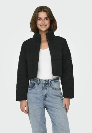 Giacca nera cropped in pile testurizzato con colletto alto e due tasche con zip, abbinata a jeans blu a vita alta.