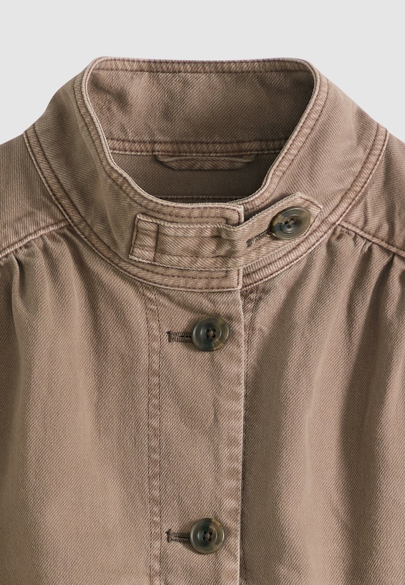 Chaqueta de algodón marrón claro con cuello alzado que incluye una correa con botón, y tres botones grandes en el centro frontal.