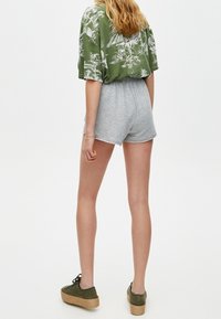 Grünes kurzärmeliges Hemd mit Palmenblattmuster, kombiniert mit grauen, hoch taillierten Shorts und olivgrünen Schuhen mit gewebter Sohle.