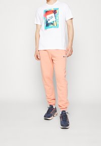 Henkilö, joka on pukeutunut valkoiseen Nike-paitaan, jossa on palmun kuva, persikanvärisiin Nike-housuihin ja sinisiin lenkkikenkään, seisoo yksinkertaista taustaa vasten.