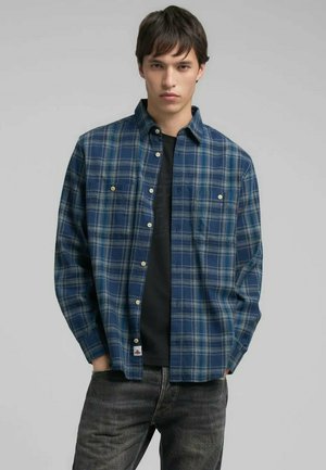 Un jeune homme portant une chemise à carreaux bleue sur un t-shirt noir, avec un jean noir délavé, se tenant les mains dans les poches devant un fond uni.