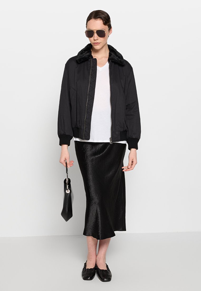 Czarna kurtka bombowca z futrzanym kołnierzem, biała koszulka, błyszcząca czarna spódnica midi, czarne baleriny i mała czarna torebka.