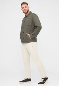 Chaqueta de botón en un verde oscuro con un bolsillo en el pecho, combinada con pantalones beige claros y zapatillas negras con cordones crema y suela color marrón claro.