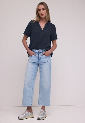 Kvinde står med hænderne i lommerne iført en mørk navy kortærmet bluse, lyse vide cropped jeans og sølv sneakers.