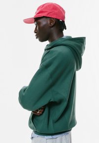 Felpa verde con tasca a marsupio e vestibilità ampia, indossata sopra una camicia a righe leggera blu e bianca. Accompagnata da un cappellino rosso.