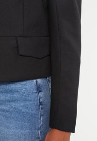 Blazer structuré noir au design élégant, tissu texturé, détail d'une seule poche et coupe ajustée, associé à un denim bleu clair.