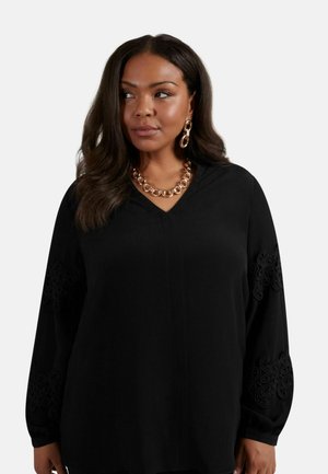 Schwarze Bluse mit langen Ärmeln, einem V-Ausschnitt und filigranen bestickten Details an den Ärmeln. Der Stoff wirkt glatt und hat eine weiche Textur.