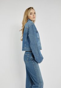 Przycięta dżinsowa kurtka w niebieskim kolorze, z długimi rękawami, klasycznym kołnierzykiem i jedną kieszenią na piersi. Teksturowany materiał z widocznymi szwami.