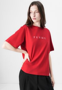 Rød t-skjorte med korte ermer laget av bomull, med ordet "FEMME" trykt i hvitt på forsiden. Enkel design med avslappet passform.