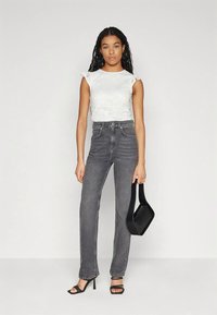 Mulher com cabelo longo trançado, usando uma blusa branca sem mangas, jeans cinzentos de cintura alta, saltos pretos e segurando uma bolsa preta.