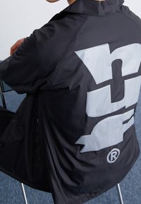 Musta tuulitakki mattapinnalla, jossa on suuri vaaleanharmaa logo selässä, ja minimalistinen muotoilu säädettävällä kauluksella.