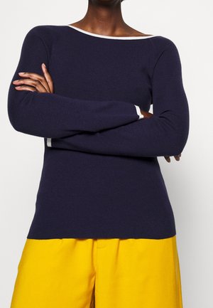 Navy blue knit sweater with a wide neckline, featuring contrasting white trim. Long sleeves and a fitted silhouette. Paired with yellow pants.